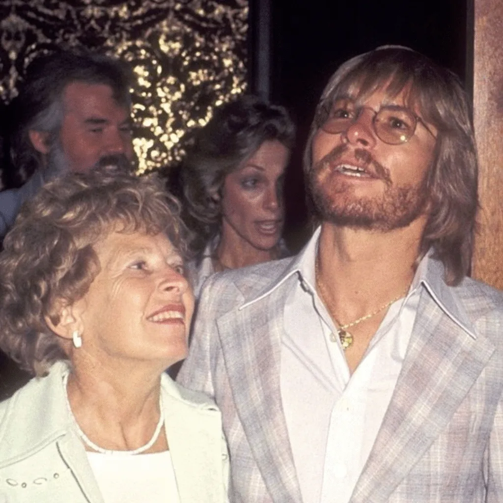 “THE NIGHT HIS MOTHER HEARD A SONG… AND REALIZED HER BOY WASN’T A BOY ANYMORE.” John Denver wrote “Sunshine On My Shoulders” on a quiet afternoon in a little cabin, the kind of day when the light feels softer and time moves slower. When he brought the demo home for his mother to hear, she sat perfectly still, her hands gently pressed together in her lap. When the last line faded, she turned her face away for just a moment — long enough to wipe her eyes. John thought he had done something wrong. But she shook her head and whispered, “You’ve grown up, John. I can’t keep you all to myself anymore.” It was the first time she understood that his music would carry him far — farther than her arms ever could. Featured image