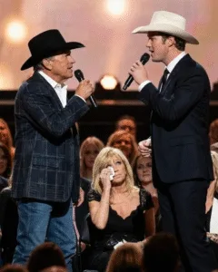 “THIS WASN’T A DUET. IT WAS A FATHER OPENING THE DOOR.” The crowd thought they were watching a concert. Then George Strait gave a small nod toward the wings. The band held the note. The lights softened. Something shifted in the air. Bubba Strait walked out, calm but steady. In that second, the King of Country wasn’t a legend anymore. He was a father inviting his son into the circle. No screaming. People stood. Quietly. Like they knew this wasn’t for applause. In the seats, Norma Strait rose too. Hand over her heart. Eyes bright. Watching the two men she loves share a song that felt lived-in, not practiced. No tricks. No shine. Just blood and time and a melody that finally found its place. And there’s more to this moment than the stage ever showed.
